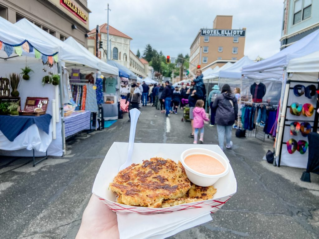 Crab cakes purchased at the Astoria Sunday Market, with tents and vendors in the background.