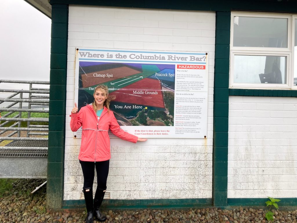 Author wearing coral pink rain jacket, smiling in front of a USCG sign about the Columbia River Bar.