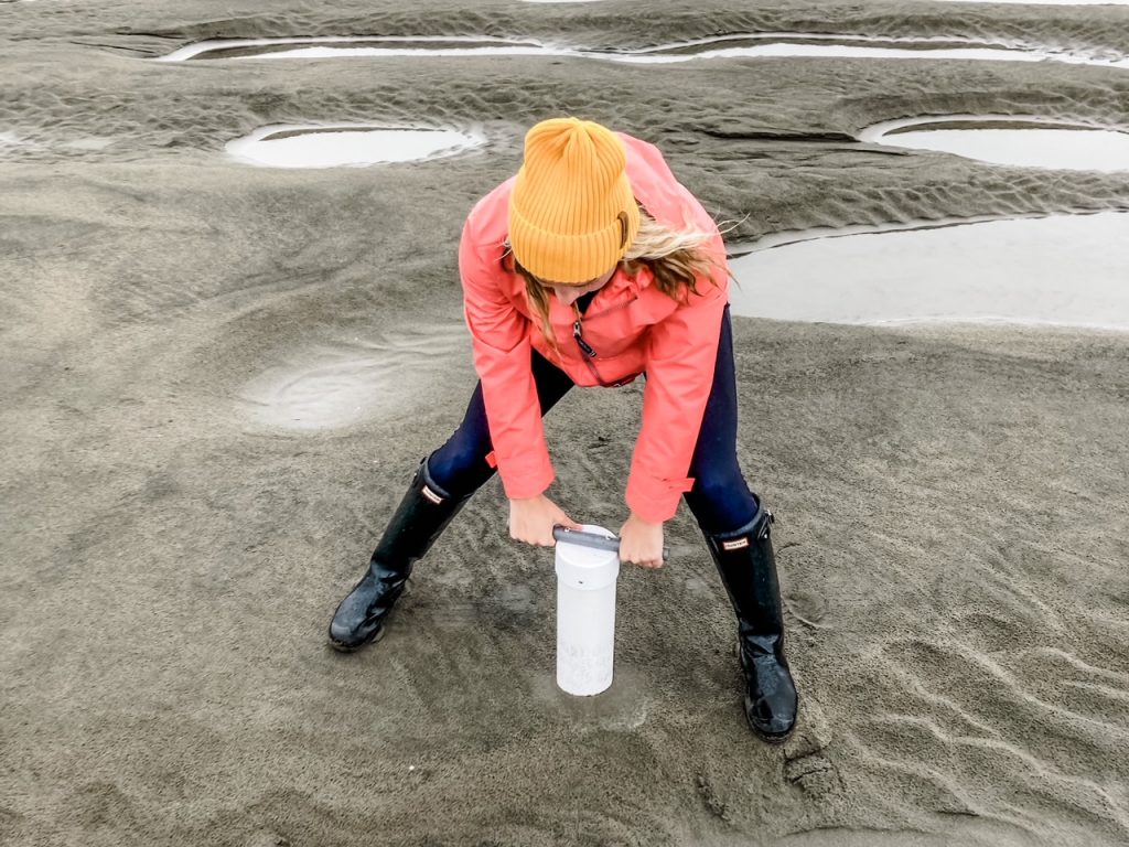 Author at the beach wearing pink rain jacket and yellow beanie, driving clam gun into the sand.