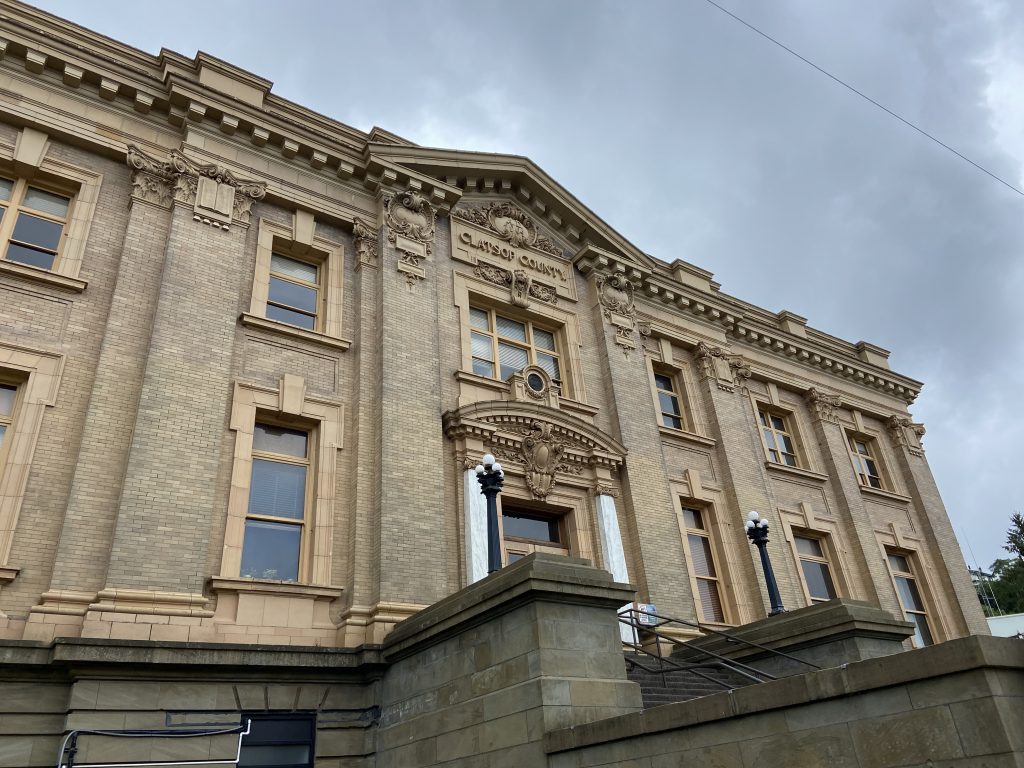 Clatsop County Courthouse, a large, beige brick building, takes up the frame against a cloudy gray sky.