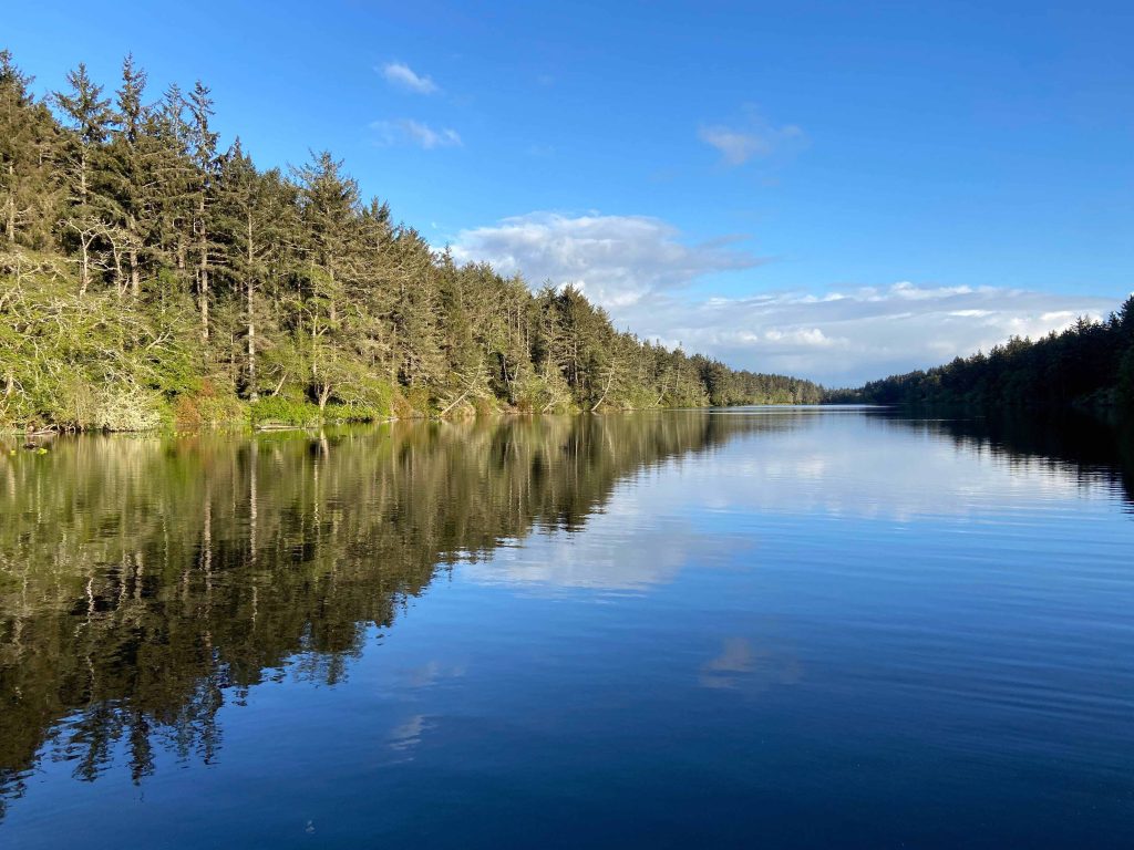 Blue sky and spruce trees are reflected in glassy Coffenbury Lake in Fort Stevens State Park.